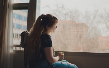 A young woman rests in a chair and looks out the window. The woman is holding a mug.